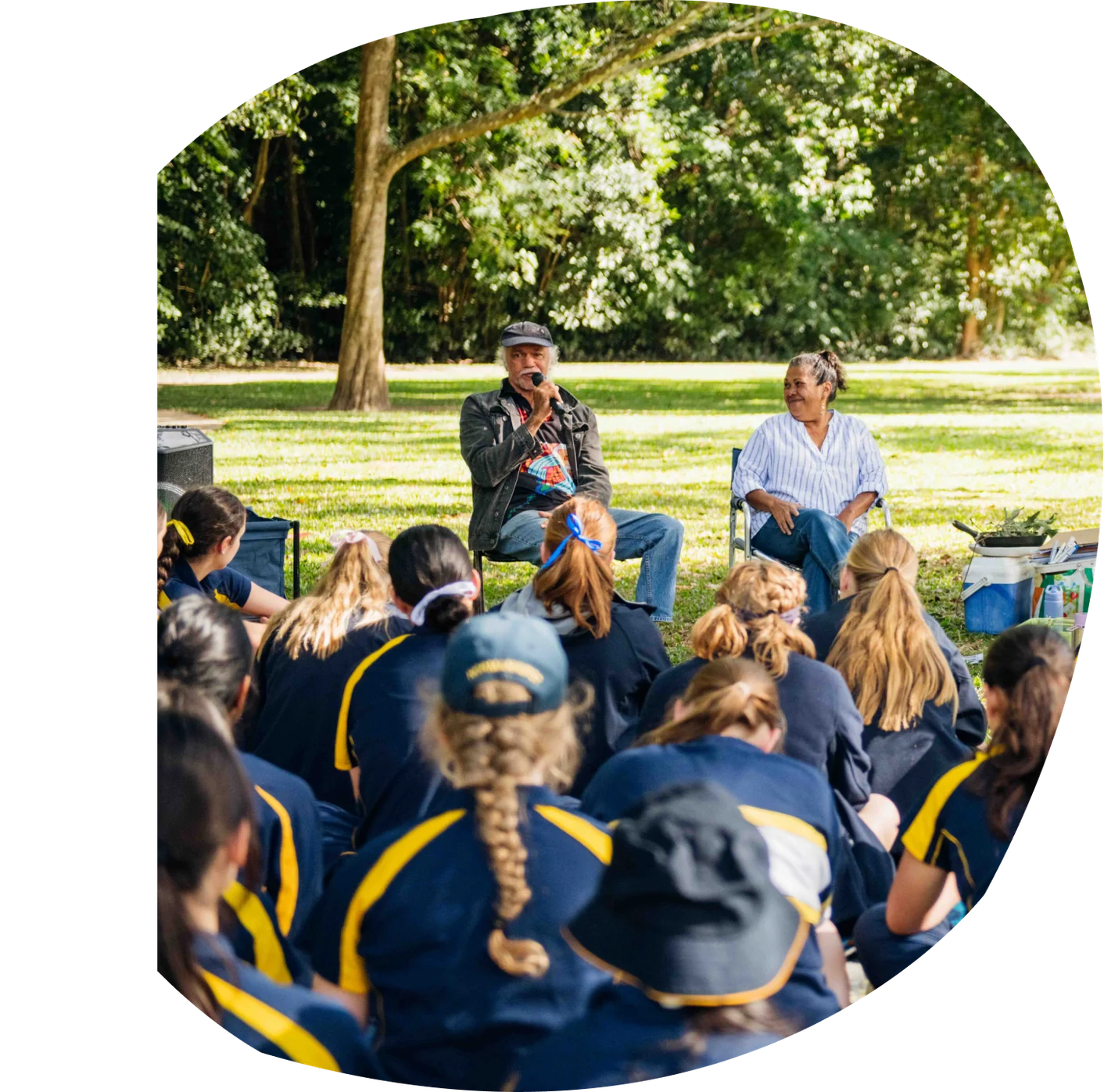 Students in school uniform sitting on the grass listening to an Elder speaking into a microphone.