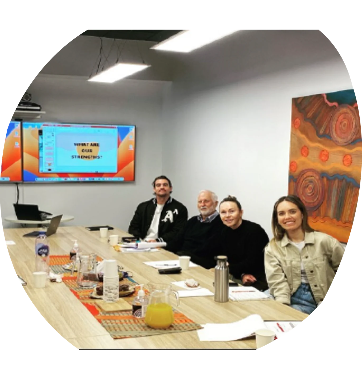 Five people seated along a meeting table smiling at the camera, with an Aboriginal artwork on the wall behind them.