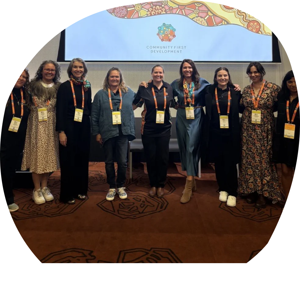 A group of women wearing conference lanyards stand in front of a Community First Development presentation screen.